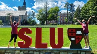 Two students stand next to large foam letters that spell out SLU on a sunny summer day. 