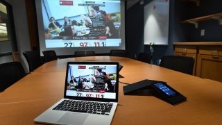 An open laptop sits upon a large conference table in a renovated library space. There's a large projector screen in the background.