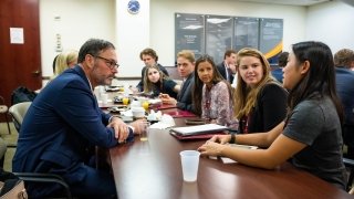 Alumni sit across from current students at a long conference table in a corporate office. A student at the head of a table is asking a question.
