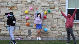 Three students stand in front of a stone wall with their arms in the air. There are colorful balloons behind them.