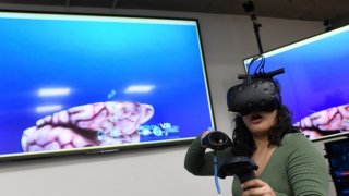 A student wearing a virtual reality headset experiments with virtual reality software. There are two large television screens behind them.