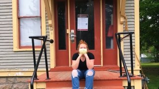 A student, wearing a mask, sits on the front steps of a brightly colored house.