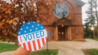 An "I Voted" sticker held up in front of an elegant stone library and fall foliage on the Saint Lawrence University campus. 
