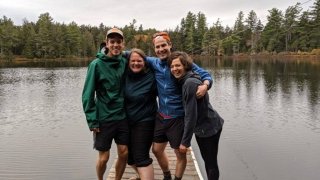 Four Saint Lawrence Adirondack Semester alumni, wearing rain jackets, stand on a dock over water in the Adirondack Park. 