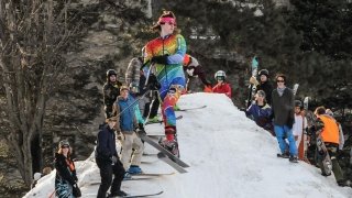 Alex Andrews, wearing a tie dye ski uniform, launches into the air off of a ski jump. A crowd of skiers and snowboarders stand on the jump behind Alex.