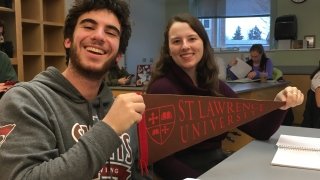 Jesse Meyer and Maddy Schumacher, holding a Saint Lawrence pennant, sit in a classroom. There are other students in the background. 