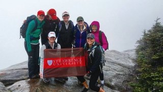 Seven Saint Lawrence students, wearing rain jackets and hats, stand on the summit of a mountain, in the rain, holding a Saint Lawrence University flag.