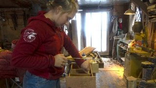 A Saint Lawrence University student, wearing a red hooded sweatshirt, saws into wood in a workshop.