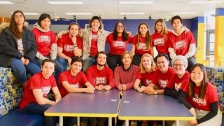 A group of students wearing red shirts sit in two rows among booths in a school cafeteria.