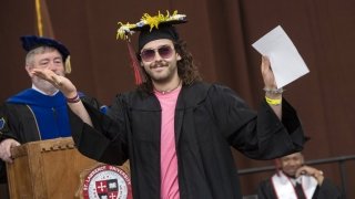 A graduate wearing sunglasses walks across the stage while waving their hands and smiling at the camera.