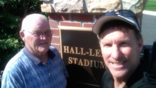 Two men stand in front of a brick and stone pillar with a plaque
