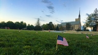 A small American flag is in the foreground sticking out of a grassy quad where numerous other small flags are placed just outside a chapel in the distance.