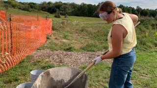 A student wearing jeans, a t-shirt, and goggles, shovels cement from a wheelbarrow. There's a large field in the background.