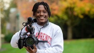 Kaleb Davis, wearing a gray Saint Lawrence sweatshirt, holds up a large video camera.