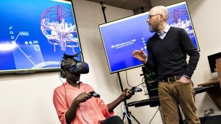 A student sits as they explore a virtual reality through a headset they are wearing as a faculty member describes the experience.