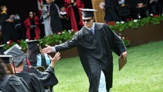 A Saint Lawrence student, wearing graduation attire and sunglasses. high-fives a fellow member of the graduating class. 