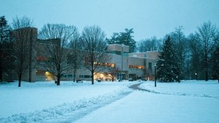 The brutalist-style library on a snowy northeastern campus at dusk. Lights from inside the library glow warmly.