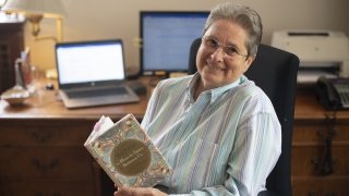A photo of Reverend Kathleen Buckley sitting and smiling in front of her desk with a book in her hands.