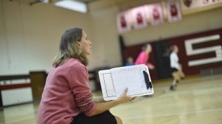 A photo of Shelly Roiger sitting on the sidelines of a basketball court with a clipboard in hand as she shouts out to players.