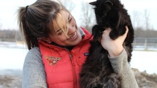 A young woman holding a black barn cat outside in the winter