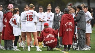 A photo of the women's lacrosse team huddled together in a meeting on the field, coach Hannah Corkery is crouching down, the back of her coat reading St Lawrence University.