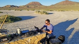 A photo of a student set up and working at a desk in the desert that has multiple devices plugged in. 