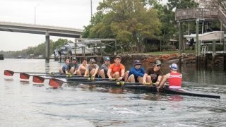 The men's crew team row together on a boat on a river in Tampa Bay on a warm, overcast day.