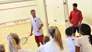 A photo of women's squash coach Scott Denne demonstrating something to the team who are sitting and watching.