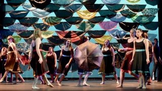 A photo of St Lawrence dancers performing on stage. They are all wearing black dresses and are dancing in front of a wall full of umbrellas.
