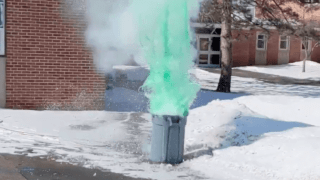 Green mist pours out of a grey garbage can sitting in the snow on a sunny day. 