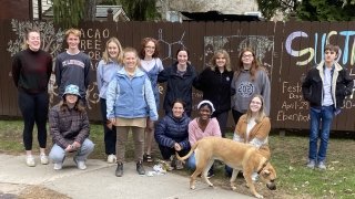 A group of students and Sara Ashpole stand in front of a brown fence that showcases their collective chalk street art project about chocolate.