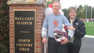 Don and Sue Martin stand in front of Hall-Leet Stadium at North Country Field on the Saint Lawrence University Campus. Don is wearing a grey Saints Lacrosse shirt and holding a lacrosse helmet and stick as a lacrosse game takes place in the background.