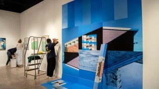 Students hang up artwork at the Richard Brush Art Gallery. There is a large piece of artwork on the white wall featuring several shades of blue.