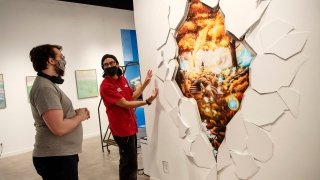 A student, wearing a red shirt, stands next to their art project and explains its inspiration to an exhibition attendee.