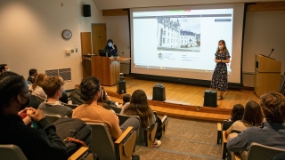 A student stands at the front of a lecture hall filled with people and presents the results of their off campus study research project. There's a large projector behind the student.