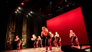 A large group of students perform on stage. There is a large red back drop behind them.