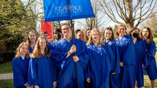 A large group of students, wearing royal blue robes, stand together on a sunny day. One student holds a banner that reads, Saint Lawrence University.