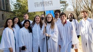 A group of students, wearing white robes, stand together on a sunny day. One student is holding a large white banner that reads, Saint Lawrence University.