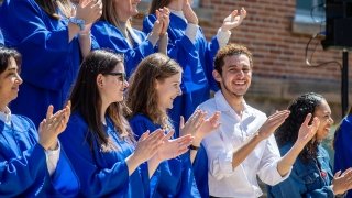 A large group of students, wearing royal blue robes, clap their hands in celebration of Moving-Up Day. On student, wearing a white shirt, smiles at the camera.