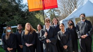 A group of students, wearing black robes, stand together on a sunny day. One student holds a large orange and red banner that reads, Saint Lawrence University.