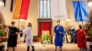 Four students stand at the front of Gunnison Memorial Chapel and hold large organge, red, white, and blue banners.