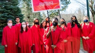 A group of students, wearing scarlet robes, stand together on a sunny day. One student holds a large banner that reads, Saint Lawrence University.