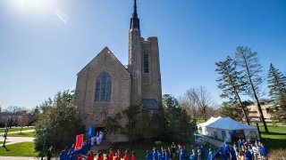 A large group of students, wearing red and blue robes, gather outside of Gunnison Memorial Chapel on a sunny day for the Moving-Up Day celebration.