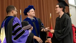 Wearing graduation regalia, Michael Shuckers hands a diploma cover to his son on the Commencement stage.