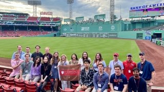 A group of students holding a Saint Lawrence University flag stand in the stands in front of the field at Fenway Park.