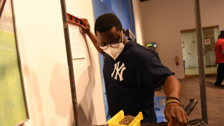 Michael Yirenkyi '22, wearing a New York Yankees t shirt, reaches for a hammer while installing his art piece on a gallery wall.