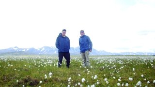 Jon Rosales and a fellow research, wearing royal blue rain jackets, stand in a field of green grass and white flowers. Large snowcapped mountains are in the distance.