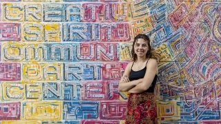 Mikayla Quinn, wearing a black tank top and patterned skirt, stands in front of a colorful mural at a community art center.