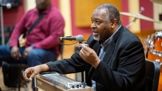 A musician sits at a keyboard and speaks to a crowd through a microphone.