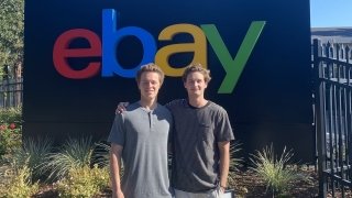 Two college-aged men wearing casual clothing stand in front of the eBay sign.